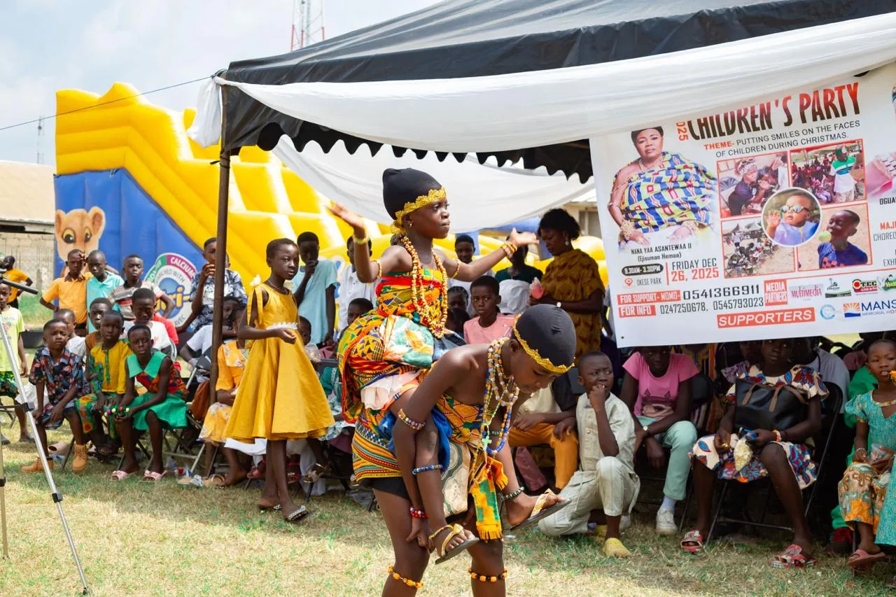 Two children in kente cloth and beads perform a cultural dance in front of seated children and the party banner.
