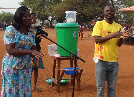 A ministry member speaking into a microphone beside a handwashing station at an outdoor gathering.